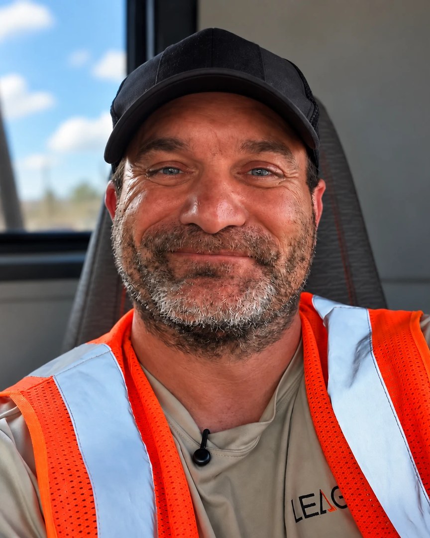 Alex, a League Trucking driver, smiling in the cab in a League shirt and safety vest.